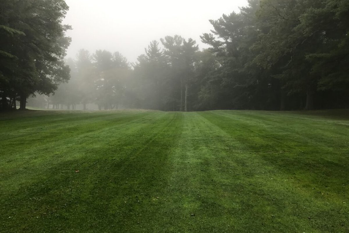 Foggy morning view of Cedar Hill Golf Club in Stoughton, MA with lush green fairway lined by tall trees and mist in the background