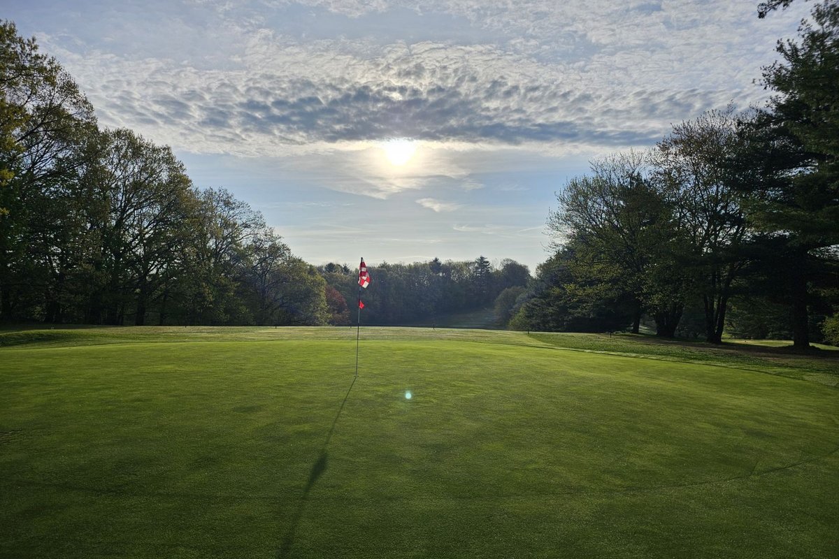 Morning sun rising over the green at Agawam Municipal GC, casting long shadows from the flagstick, surrounded by trees.