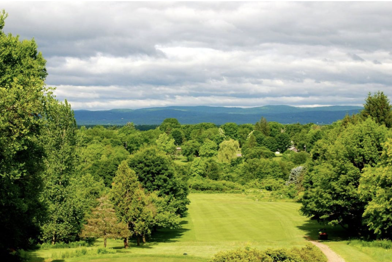 Wide fairway at Cherry Hill Golf Course in Amherst, MA, bordered by lush green trees under a cloudy sky with distant rolling hills