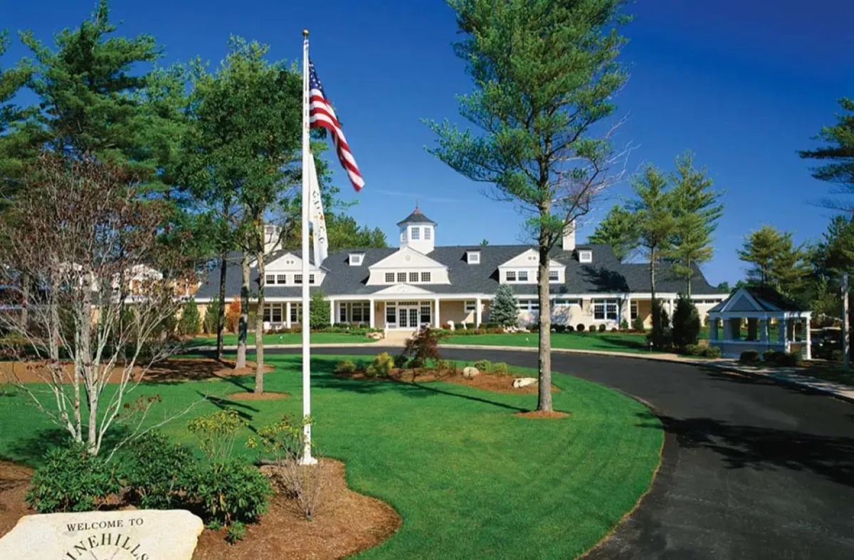 Clubhouse at Pinehills Golf Club in Plymouth with manicured lawns, American flag, trees, blue sky, and welcome stone in foreground