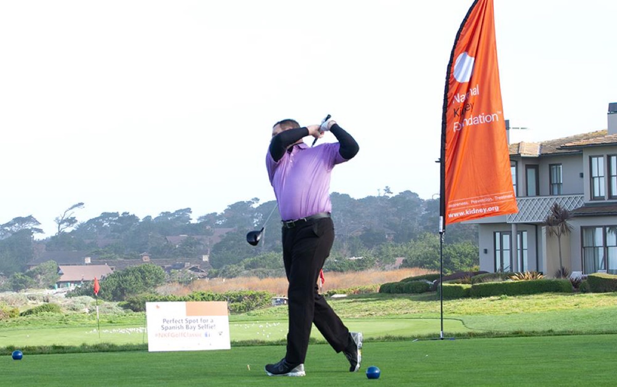 Golfer in purple shirt finishes swing on TPC Boston tee box, National Kidney Foundation flag and sponsor sign nearby, clubhouse in background